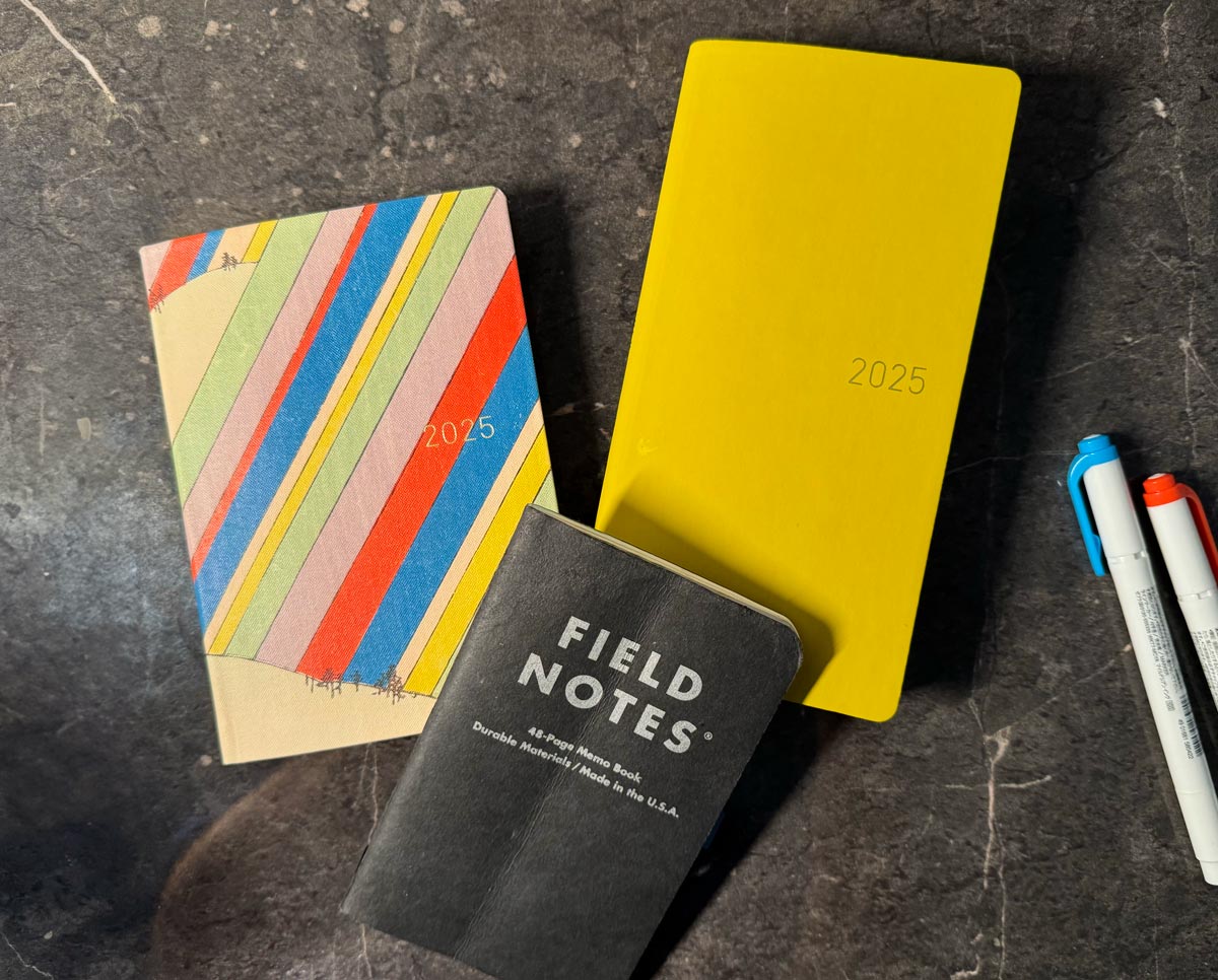 Three books on a desk: A 2025 HON with Rainbow cover, a bright yellow Hobonichi Weeks, a small black Field Notes notebook.